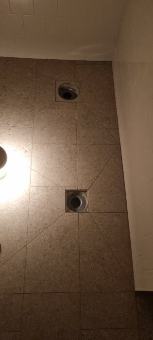 Två avloppsbrunnar i ett badrum med grått klinkergolv och vita väggar i bakgrunden.