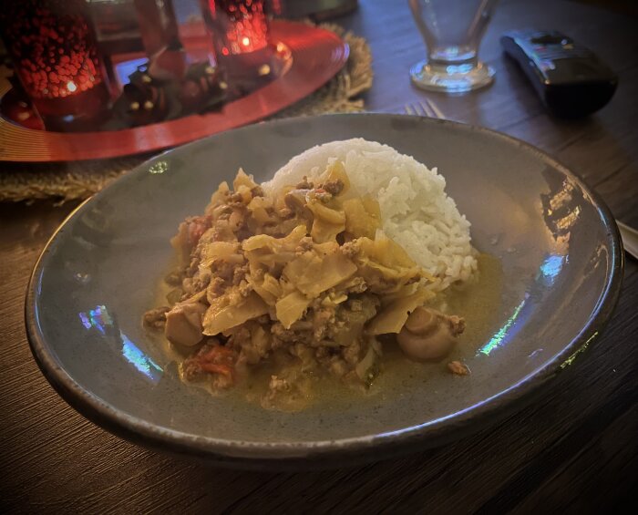 Ris och köttgryta med kål och svamp på tallrik, serverad på ett dukat bord. Ljusstakar och dekor i bakgrunden skapar stämning.