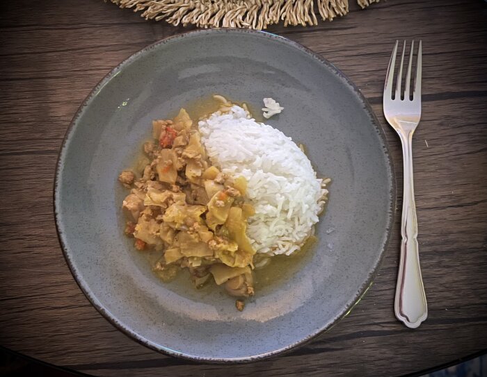Tallrik med ris och gryta, bestående av köttfärs och grönsaker, på ett mörkt träbord med en gaffel bredvid.