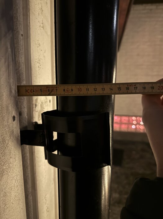 Mätsticka visar avståndet mellan ett svart rör och en fasad, cirka några centimeter, med en hand som håller i stickan.