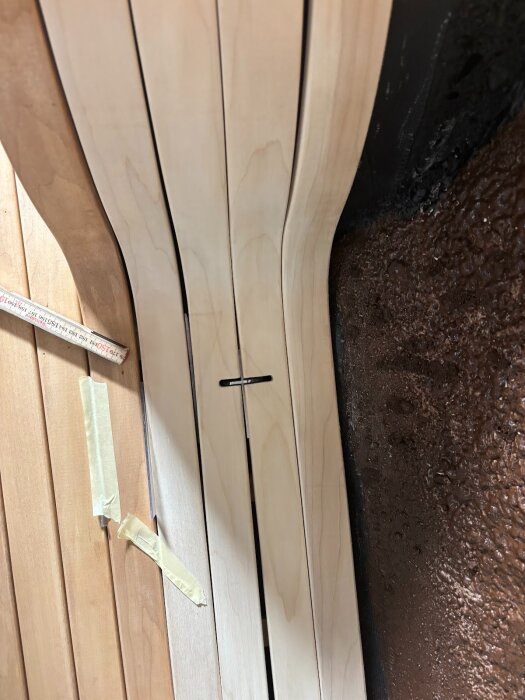 Närbild på träpanel med tunna ribbor i ljus och mörk träfärg, monterade mot en svart yta med maskeringstejp fäst på.