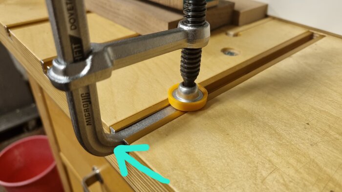En metallklämma med Microjig-logotypen monterad i en T-track-profil på en arbetsbänk, med en pil som markerar passningen.