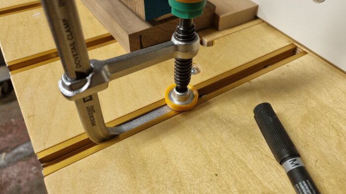 En Microjig dovetail-klämma fäst mot ett träbord med en metallspårprofil. Bredvid ligger en svart markeringspenna.
