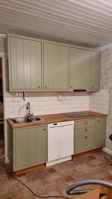 Nymålat kök med grönblå skåpluckor och träbänkskiva. Vitt kakel ovanför diskbänk, golv i bruna toner. Diskho och diskmaskin syns.