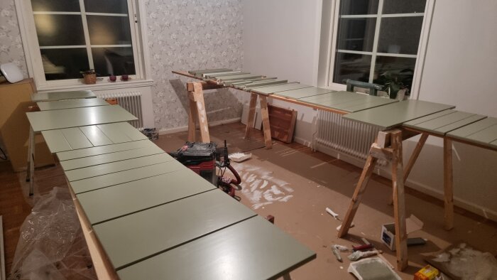 Nymålade köksluckor i grönt ligger på tork på bockar i ett rum med fönster och tapetserade väggar.