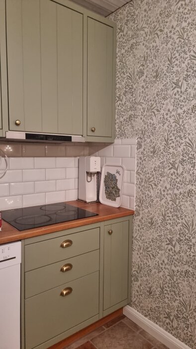 Nymålat kök med grönfärgade skåpluckor, vit kakelvägg och blommönstrad tapet, sett ovanför en diskbänk med kaffemaskin och bricka.