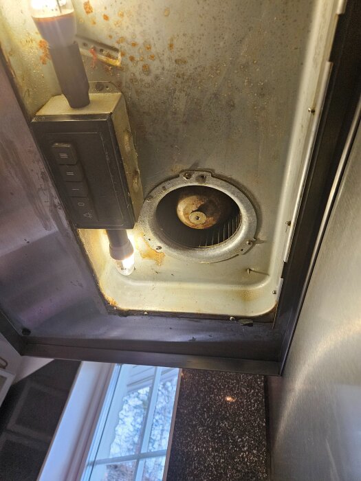 Fläktkåpa i kök med smuts och rostfläckar samt belysning. Kontrollpanel med knappar på vänster sida. Fönster syns i bakgrunden.