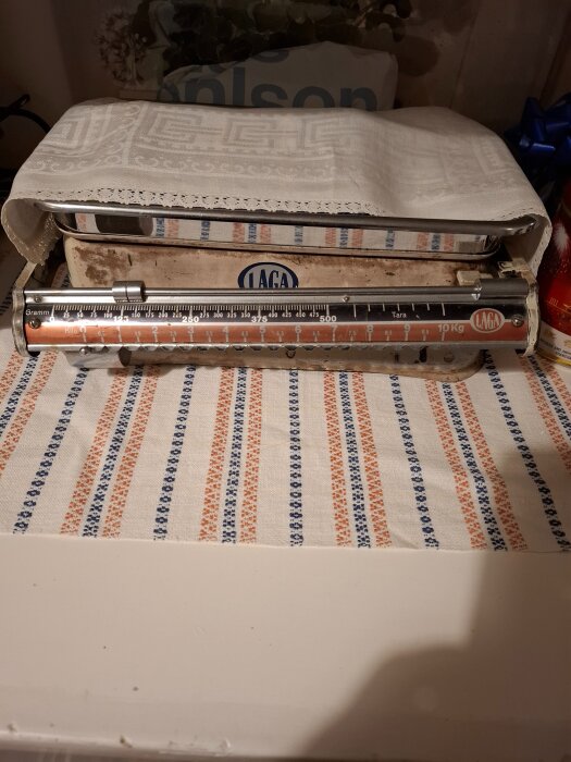 Gammal mekanisk köksvåg med LAGA-logga, täckt med vit duk, står på en randig bordsduk i blått och orange.
