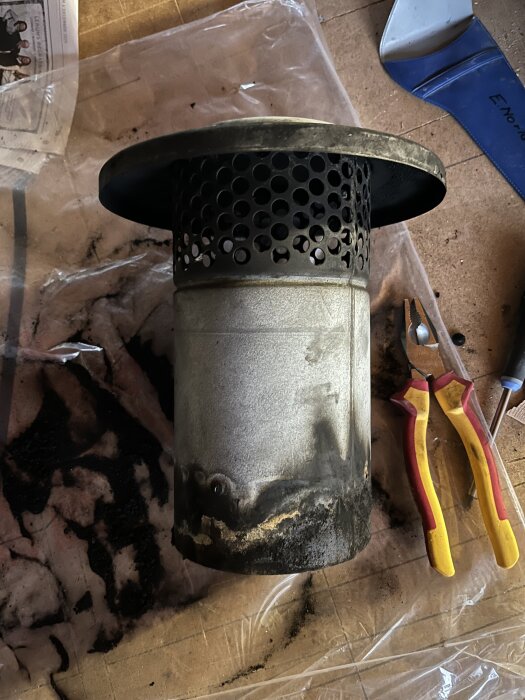 Rund, metallisk skorstensavslutning med perforerad sektion, placerad på ett bord med verktyg i närheten.