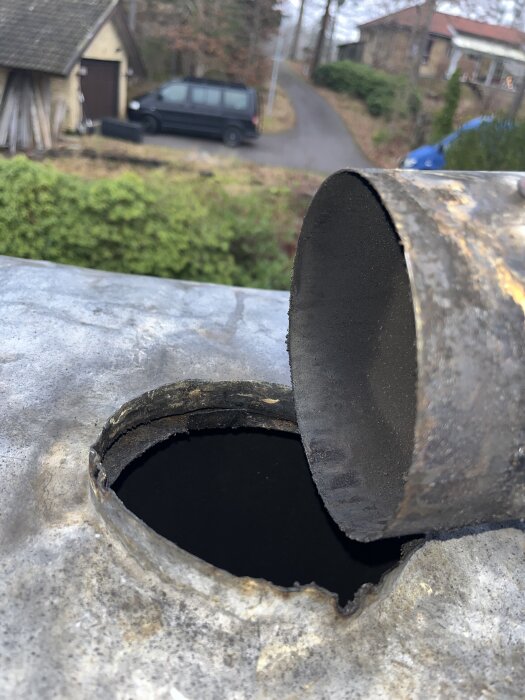 Rostig, rund skorstensavslut bredvid ett runt hål på ett tak, med naturscen och hus i bakgrunden.