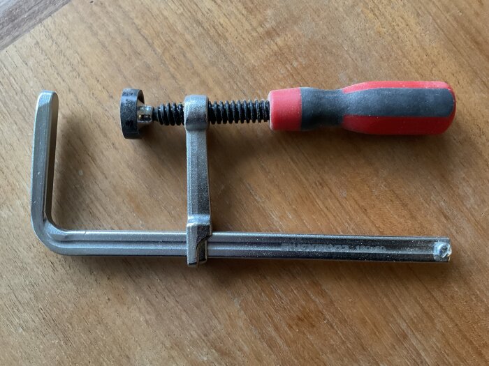 Metallklämma med röd-svart skruvhandtag på träbord.
