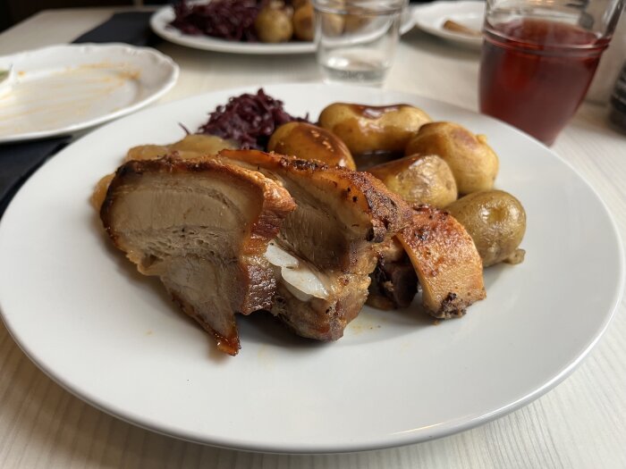 Fläskestek och revbenspjäll med ugnstekt potatis, rödkål och sås på en tallrik, serverad med ett glas dryck i bakgrunden.