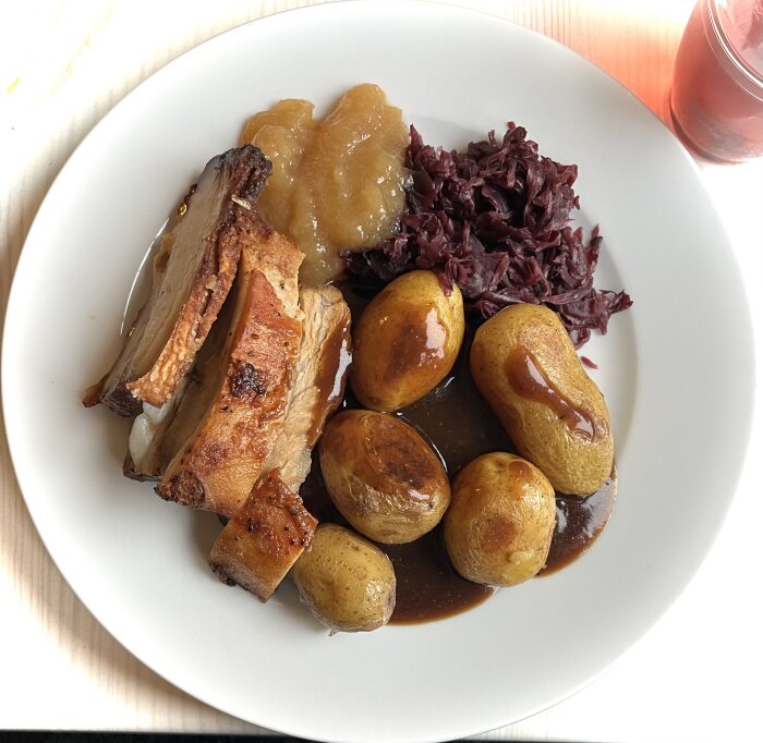 Tallrik med fläskestek, revbenspjäll, ugnstekt potatis, rödkål, sås och äppelmos.