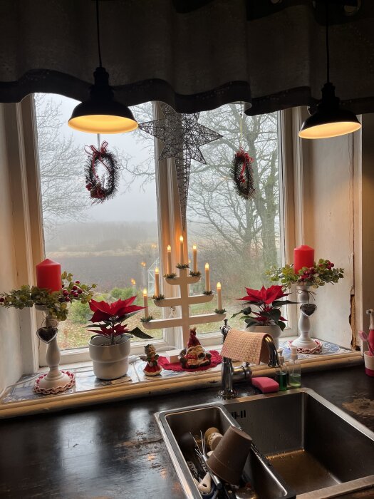 Juldekorerat köksfönster med adventsljusstake, röda ljus, julkransar och julstjärnor i krukor. Diskho och köksbänk i förgrunden.