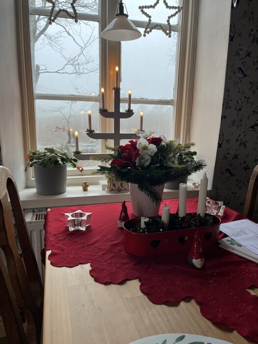 Matbord med röd julduk, adventsljusstake och juldekorationer vid ett fönster med utsikt över vinterlandskap.