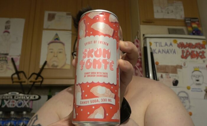 Person håller en burk "Skum Tomte" godisläsk, 330 ml. Burken är röd med vitt och har bilder av skumtomtar. Bakgrund med köksskåp och teckningar.