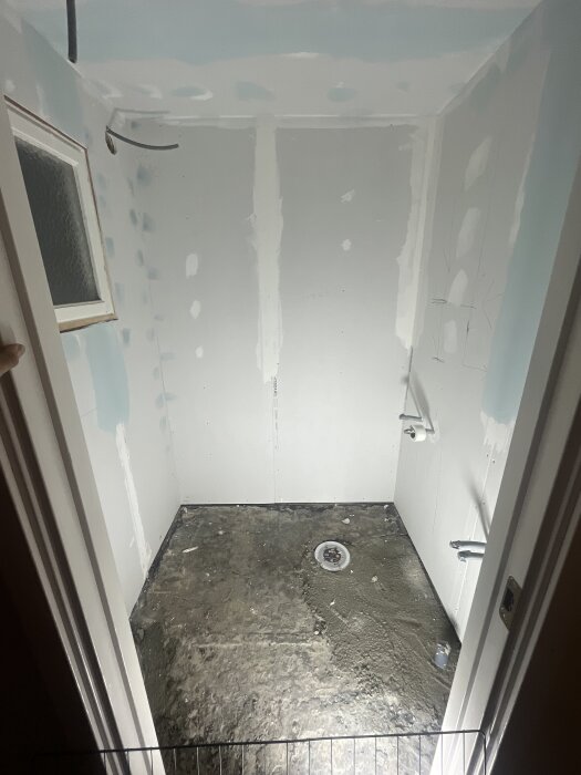 Badrum under renovering med ett gammalt fönster i våtzon 1, oisolerade väggar och synligt avlopp i betonggolv.