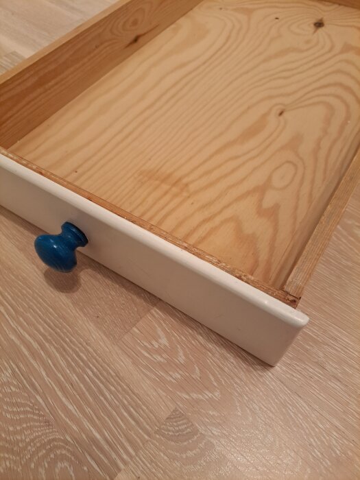 En kökslåda av trä med en vit framsida och blått handtag. Lådan är utdragen och visar träets ådring. Den står på ett ljust trägolv.