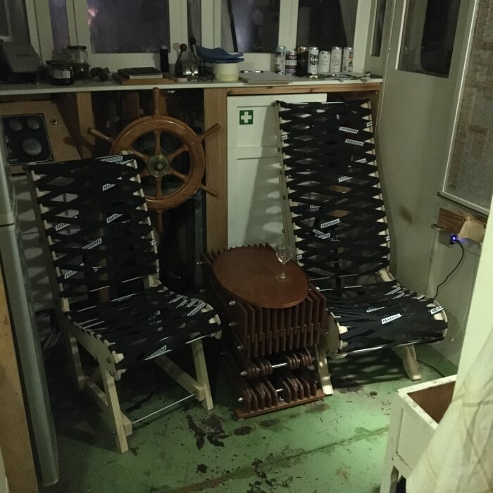 Två fällbara stolar med svart vävrygg och träben står inomhus bredvid ett bord i form av en båtstyrning och en ratt.