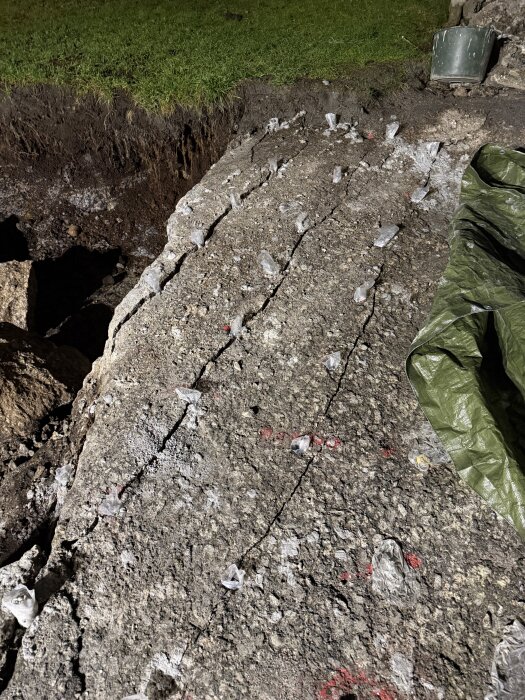 Sprucken betongyta med små plastkapslar i sprickorna, grönt gräs och plast presenning synliga i bakgrunden.