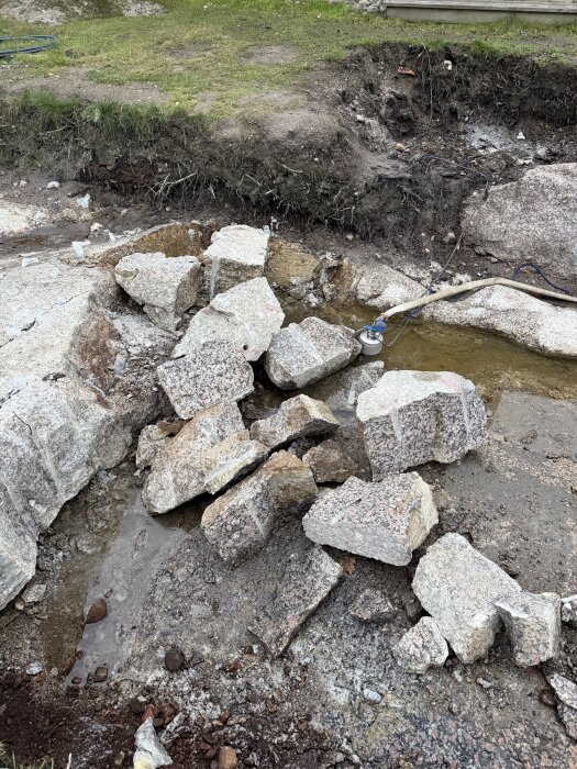 Stora stenar och jordhög vid en grävd grop, med en vattenpump och slang synlig. Gräs och jord omger arbetsområdet.