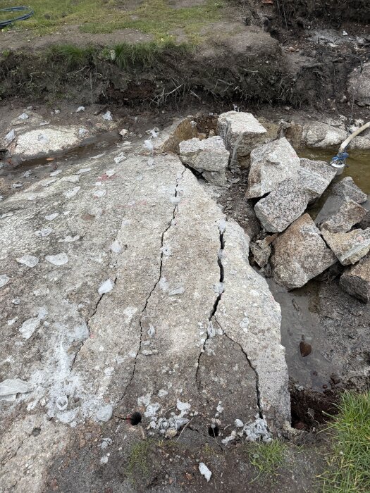 Krossad betongplatta med stora sprickor och bitar av cement vid en gräsmatta, där vatten syns vid sidan av det skadade området.