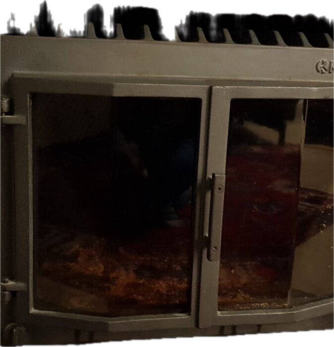 Gjutjärnskamin med glasdörrar, stängd och ej i bruk, i ett rum med röd matta i bakgrunden.