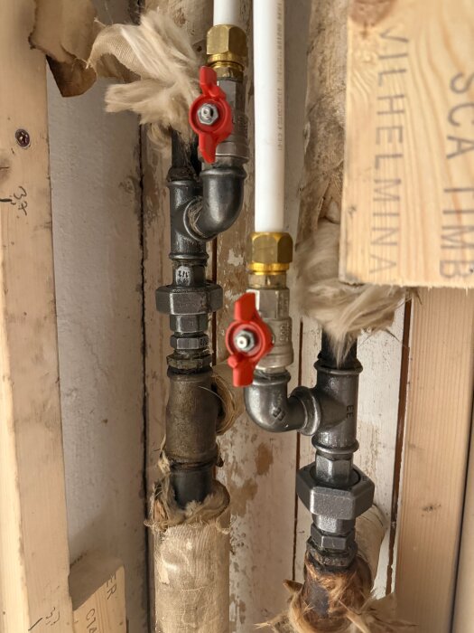 Två isolerade rör med klämringskopplingar och röda ventilhandtag i ett källarprojekt för att ansluta radiatorer.