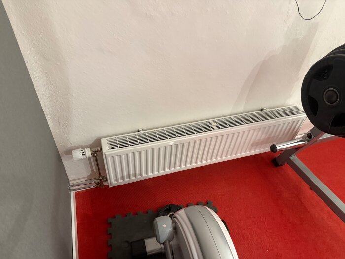 Vit radiator installerad i hörnet av ett rum med grå och vitvägg. Röd matta och gymutrustning syns på golvet.
