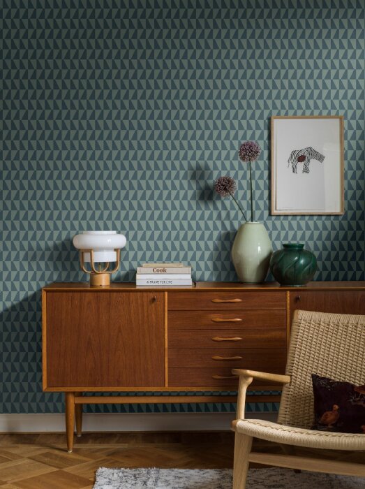 Midcentury sideboard i teak med vaser, böcker och en lampa ovanpå, framför en blå geometrisk tapet.