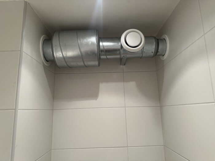 Ventilationskanal i badrum med metallrör och dämpare installerad, fästad på kakelvägg.