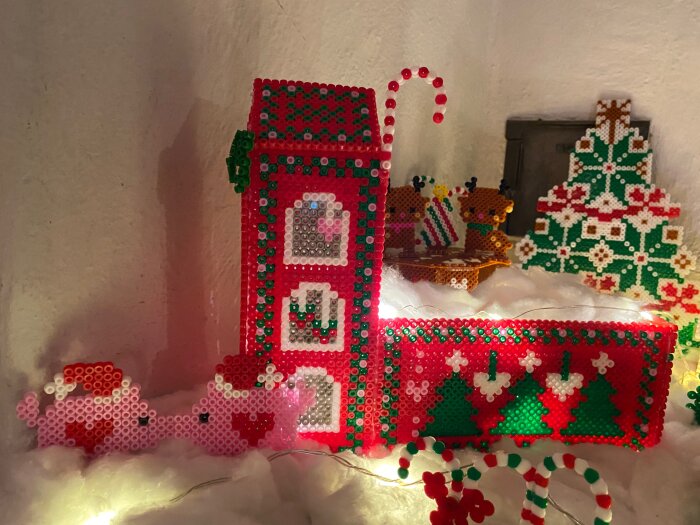 Pärlplattor med julmotiv, inklusive ett rött hus med gröna julgranar, renar och en julgran i bakgrunden dekorerar en vintrig scen.