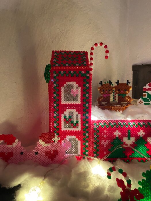 Pärlplattebyggnad med juldekor, inklusive pepparkakshus, tomtar och renar i färgerna rött, grönt och vitt, omgiven av bomull och ljusslinga.