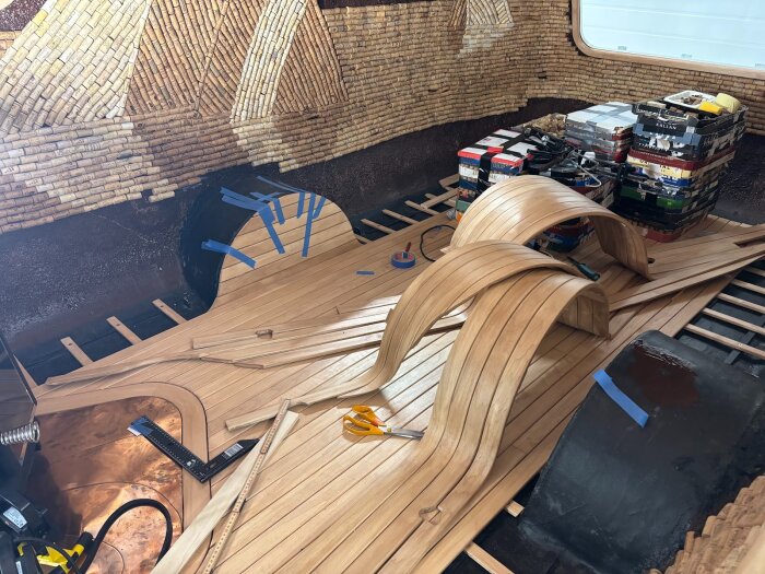 Trägolv under konstruktion med böjda plankor, verktyg och material i ett rum med korkväggar, fokus på detaljerad installation.