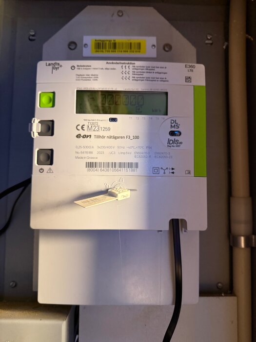 Digital elmätningsenhet med display och knappar, installerad på en vägg med synliga kablar.