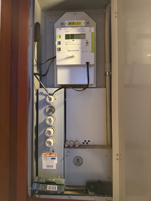 Elskåp med elmätare och huvudsäkringar. Flera säkringar syns monterade i panelen och några ligger på en hylla i botten av skåpet.
