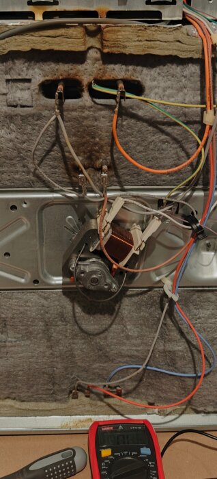 Elektriska komponenter och ledningar på baksidan av en ugn; en multimeter och skruvmejsel syns i förgrunden.