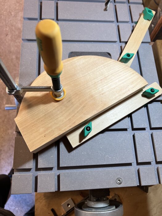 En träbit i frästa spår hålls fast med en tving på arbetsbord. Grått bord med rutmönster, gröna detaljer på träbiten.
