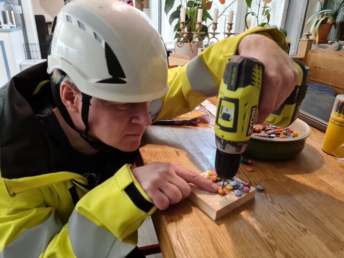 Person i hjälm och gul jacka använder en skruvdragare för att fästa godisbitar på ett träplatta, en del av ett pepparkakshusprojekt.