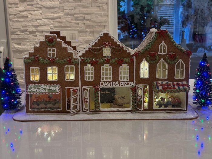 Pepparkakshus med tre butiker: Donnas bakverk, Davids pizza, och Daniel och Dianas godisbutik, dekorerat med kristyr och små röda detaljer.