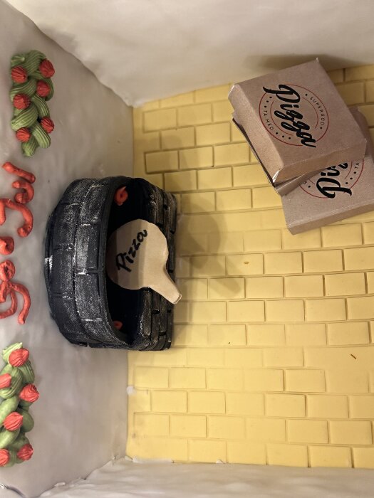 Pepparkakshusets interiör med pizzaugn och "Pizza"-spade, omgiven av små kartonger med texten "Digga" på ett gult kakelmönstrat golv.