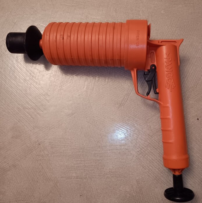 Orange lufttrycksdriven avloppsrensare liggande på ett golv, avsedd att lossa stopp i avlopp genom kraftiga luftstötar.