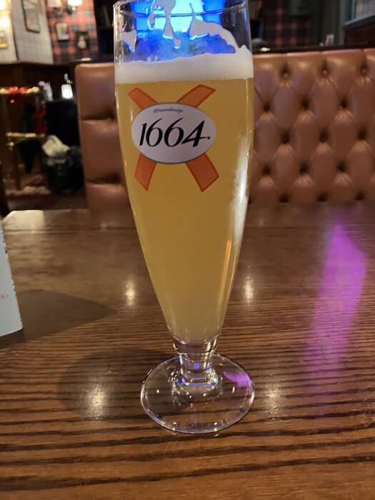 Ett glas öl av märket 1664 står på ett träbord i en pubmiljö.