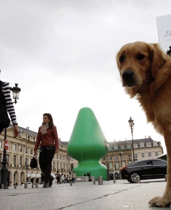 Hund framför stor grön skulptur på ett torg, liknande en julgran, med förbipasserande människor och bilar i bakgrunden.