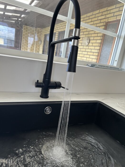 Svart kökskran med vattenstråle i en svart diskho framför ett fönster med utsikt över tegelvägg och tak.