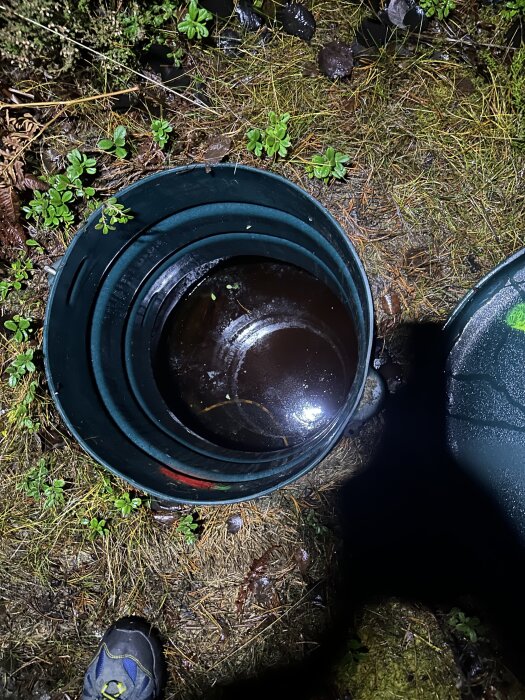 En brunn med stående vatten, omgiven av gräs och växtlighet, sedd från ovan i mörker med en fot synlig i bildens nedre kant.