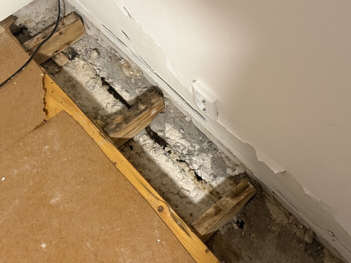 Öppet golv i gillestuga visar exponerade reglar och skadat material vid renovering från 60-talet, intill en eluttag på väggen.