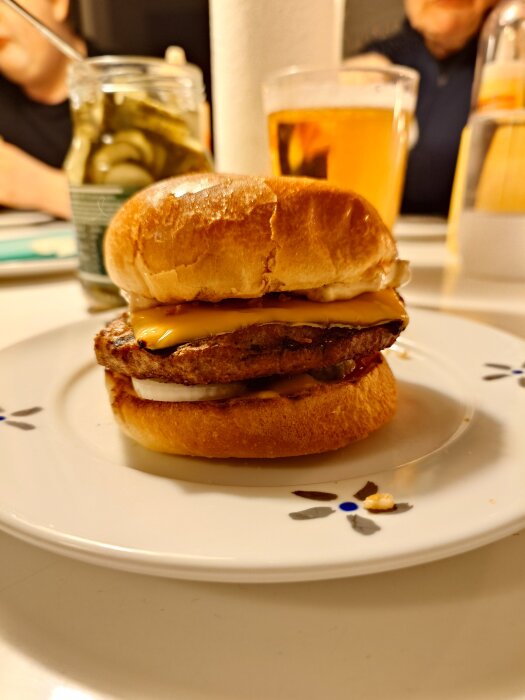 Hemgjord hamburgare med ost på ett vitt fat, en glasburk med pickles och ett glas öl i bakgrunden.