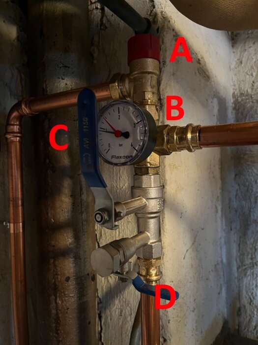 Expansionskärl med etiketterade kranar (A, C, D, E) och manometer (B). Mätsystem monterat på kopparrör i en källare.