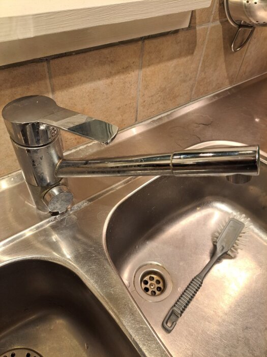 Köksblandare i rostfritt stål över en dubbelho i diskbänk, bredvid en diskborste.